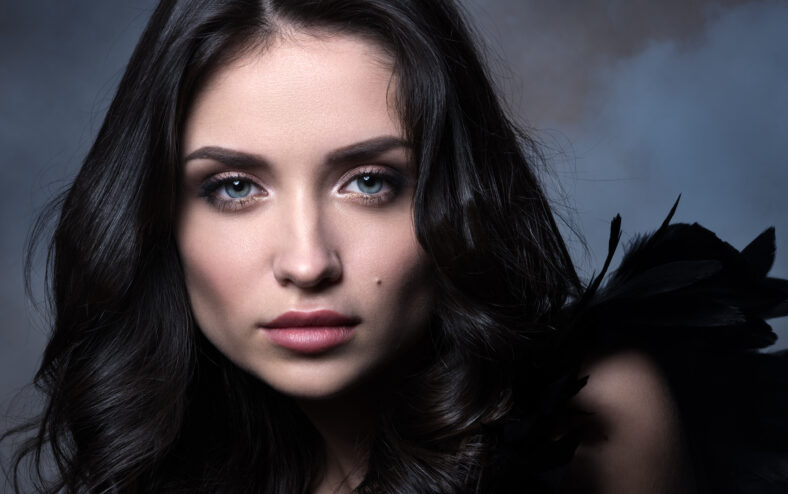 Closeup beauty portrait in cool tones. Beautiful young woman in a dark way in the cloud of smoke. Professional makeup and hairstyle. Brunette with blue eyes. Black feathers and shadows