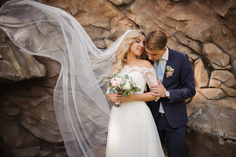 Portrait of a young bridegroom embracing a lovely blonde bride holding a bouquet of flowers on the background of wild stone