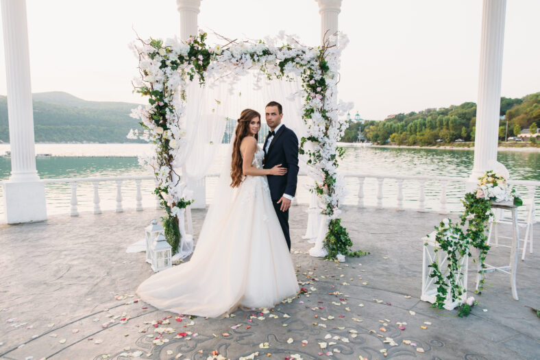 Attractive groom and bride at wedding day ceremony with arch and lake on background stands together. Beautiful newlyweds, young woman in white dress and long hairs, men in black suit. Happy family concept.