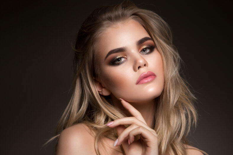 Glamour portrait of beautiful girl model with makeup and romantic wavy hairstyle. Fashion shiny highlighter on skin, gloss lips make-up and dark eyebrows.