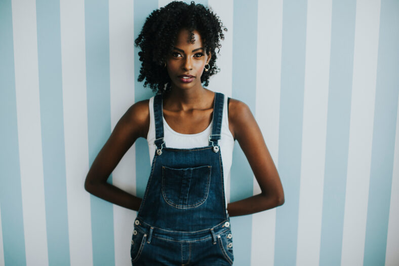Pretty young african american woman posing in jeans trousers by the wall in the room