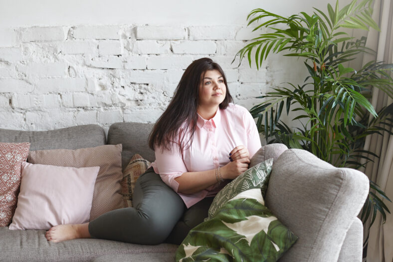 Picture of thoughtful attractive young Caucasian overweight female dressed in skinny jeans and pink shirt daydreaming at home, sitting on comfortable sofa at window, looking away with pensive smile