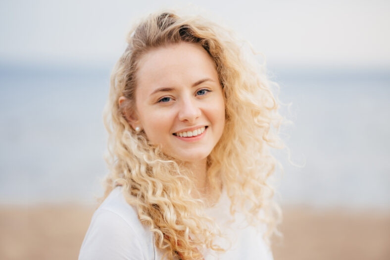 Cheerful optimistic young female has shining smile, curly hair, breathes fresh air at seaside, stands alone, has fresh skin. Attractive woman enjoys leisure time, expresses happiness. Emotions concept