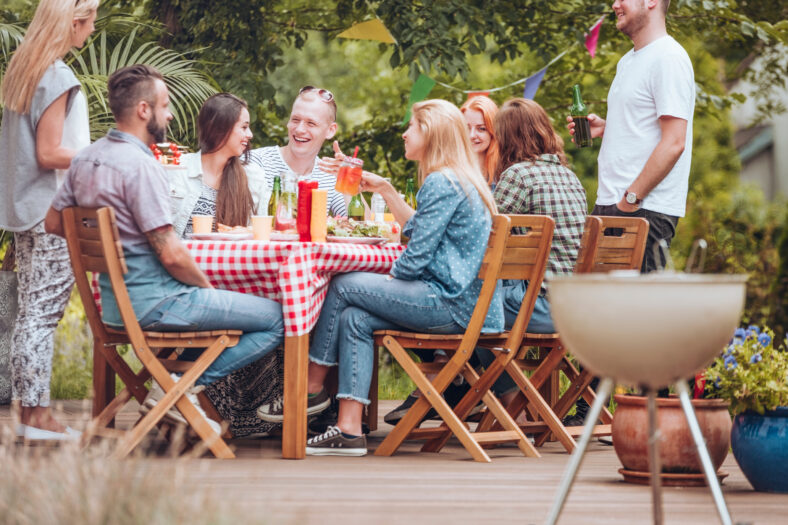 La gente se reunió alrededor de una mesa de madera, comiendo, bebiendo y divirtiéndose durante una fiesta de parrilla en la terraza.