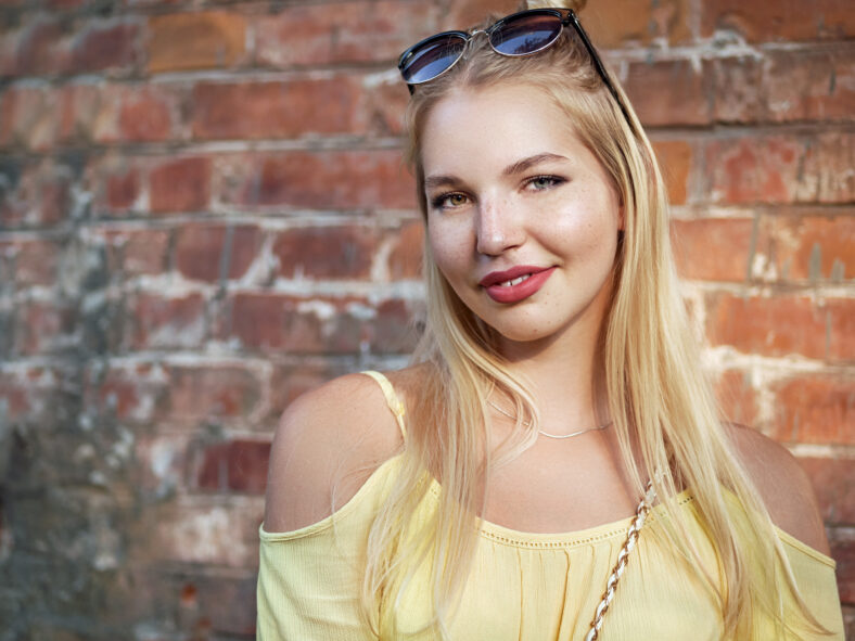 Close up portrait of young beautiful blonde woman with high bun hairstyle jeans shorts yellow blouse enjoying bright sunlight posing against brick wall