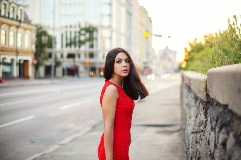 Beautiful girl in a red dress is standing near the roadway of the city, looking back.
