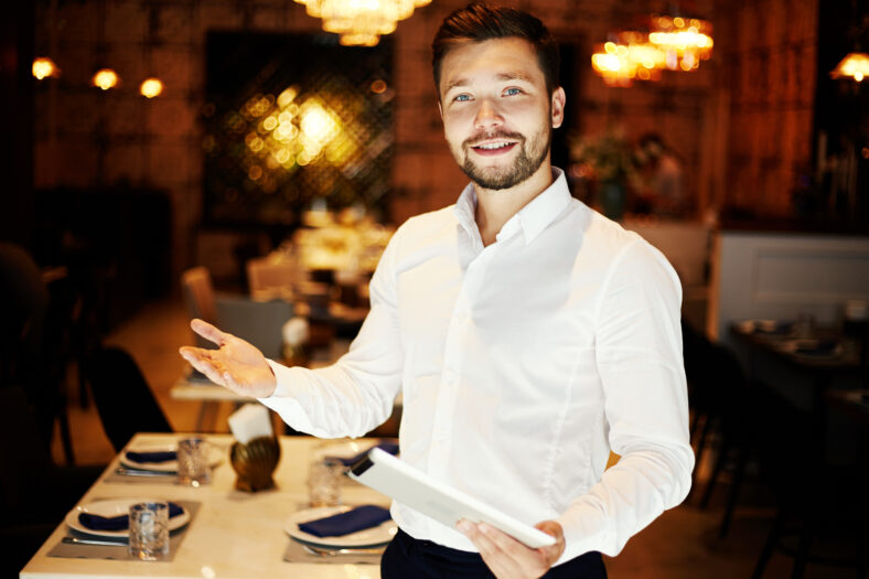 Handsome young friendly man in white shirt holding tablet and greeting with gesture smiling at camera in restaurant