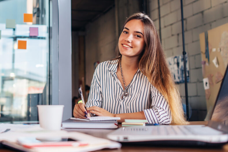 Portrait of smiling female student writing looking at camera sitting at table.
