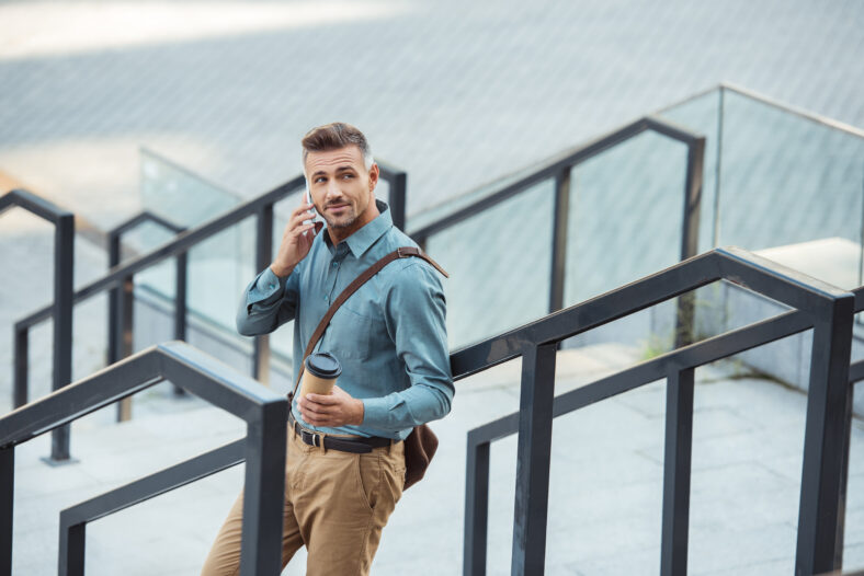 smiling middle aged man talking by smartphone hand holding paper cup while standing on stairs