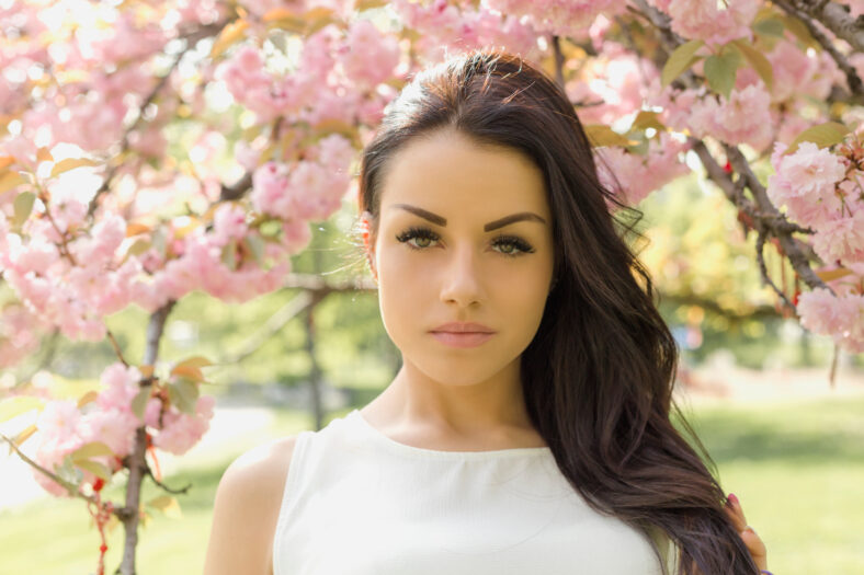 Attractive brunette woman in light dress looking at camera while posing near flowering tree