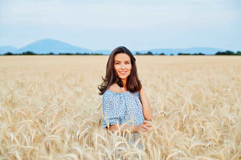 The beautiful young girl with long brunette hairs poses in the field with wheat, sunset light, turns, smiles, flirts,  happy, blue dress, France, Provence