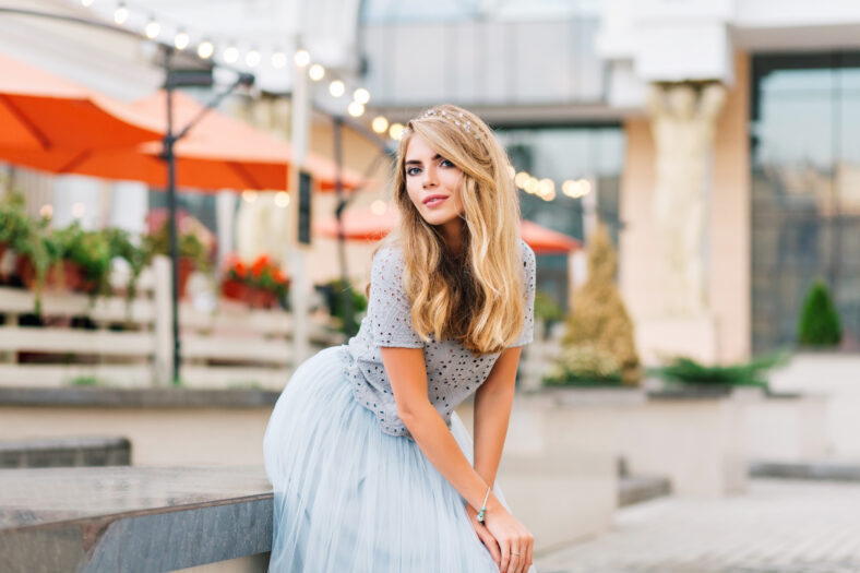 Attractive girl girl with long blonde hair in blue tulle skirt leaning on concrete bench on street. She is looking to camera