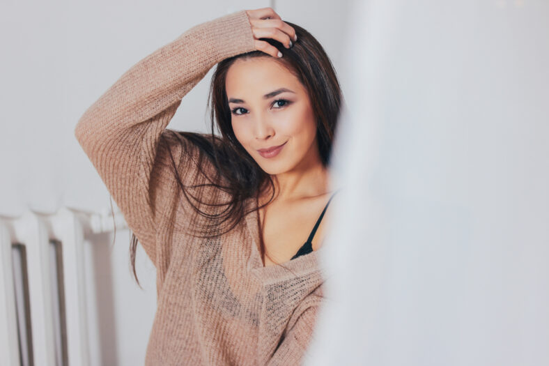 Candid portrait of smiling asian girl young woman with dark long hair in beige sweater on white curtain background