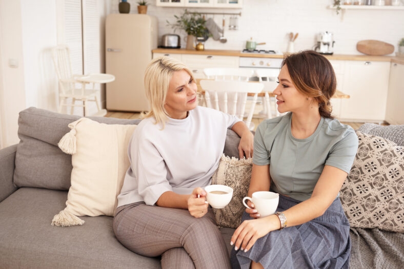 People, lifestyle, family and relationships concept. Pretty girl visiting her middle aged mother. Beautiful blonde woman speaking to her young daughter, sitting in living room with mugs, drinking tea