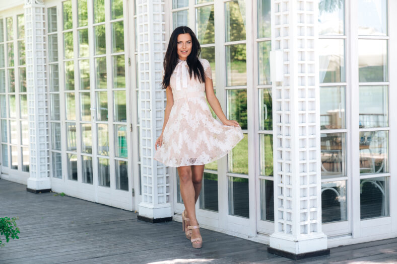 Cute brunette with beautiful long legs posing in beige colour dress outside near a wooden and glass wall.