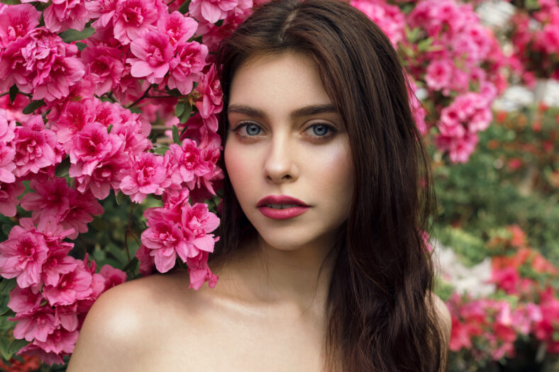 Close up potrait of beautiful young brunette woman with red lips and healthy skin with pink flowers near her face. Beauty and flowers concept