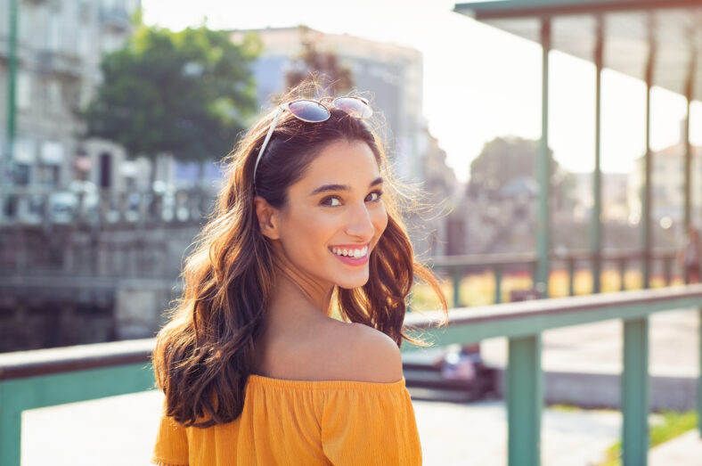 Happy latin woman looking behind while walking on street. Beautiful young woman enjoying vacation during summer. Cheerful girl in smart casual walking and looking at camera while smiling.