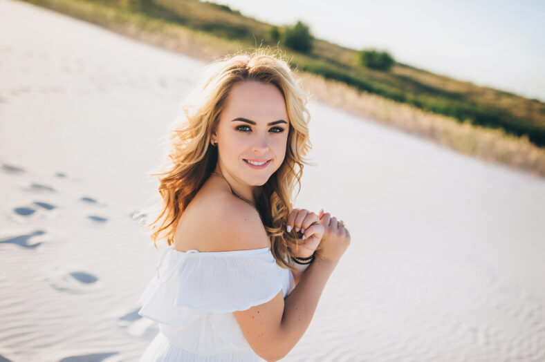A beautiful young blonde girl walks along the white sand on the beach and holds himself hand in hair and smiling. The concept of women's health and youth.