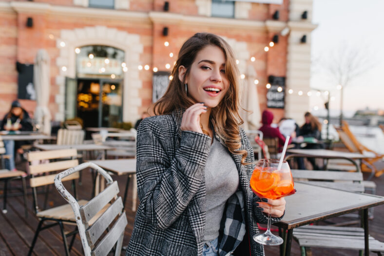 Romantic girl in casual clothes chilling in cafe in autumn weekend and tasting cocktail. Portrait of lovely european woman waiting someone in restaraunt and drinks fruit beverage.