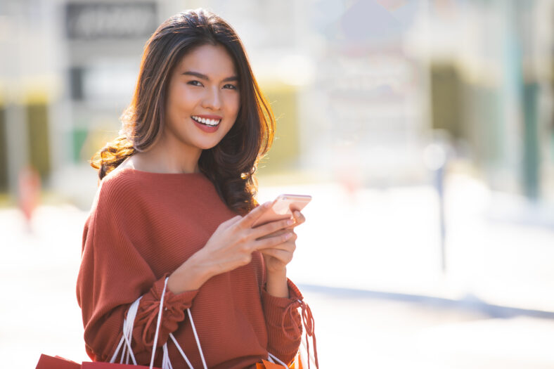 Portrait of a happy Asian pretty girl holding shopping bags while using smartphone in shopping mall background concept.