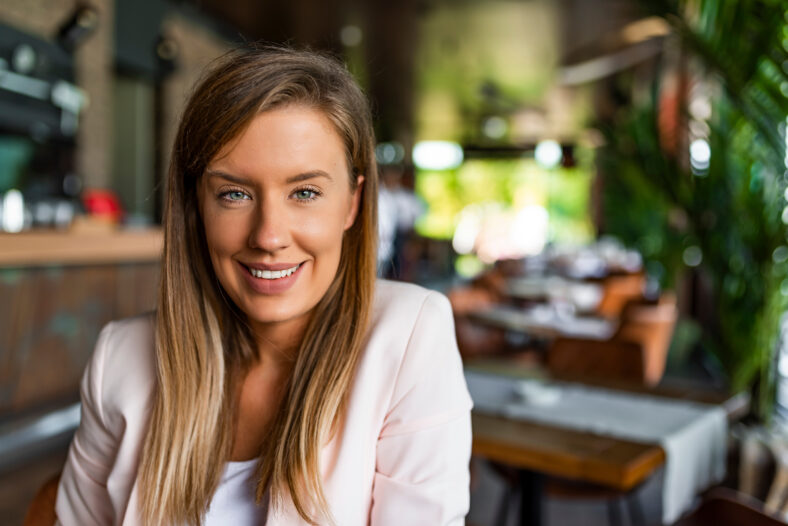 Beautiful business lady is looking at camera and smiling while working. Young businesswoman sitting at desk and working. Smiling and looking back at camera. Casual business woman looking happy and smiling