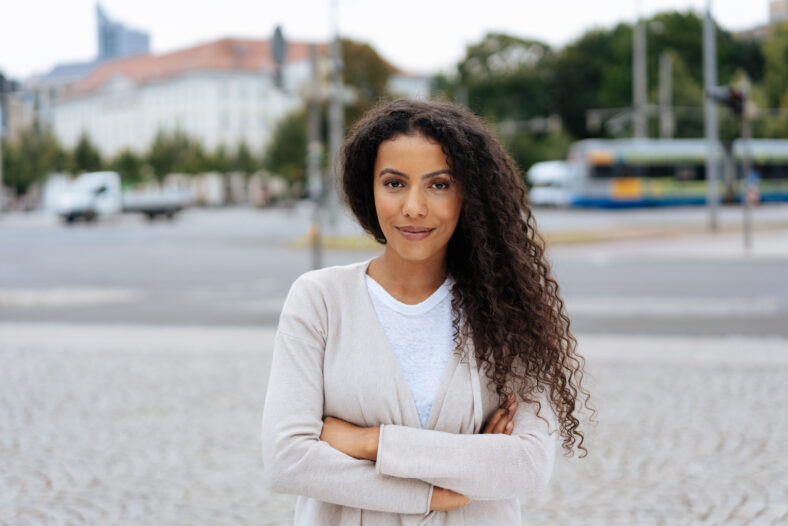 Confident young woman with a friendly smile standing in a quiet city street with folded arms looking at the camera, with copy space