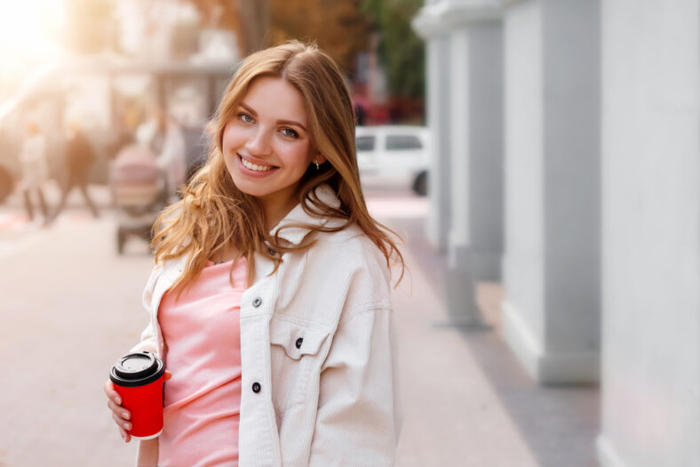 Linda chica rubia camina por la ciudad con una taza de café y sonriendo