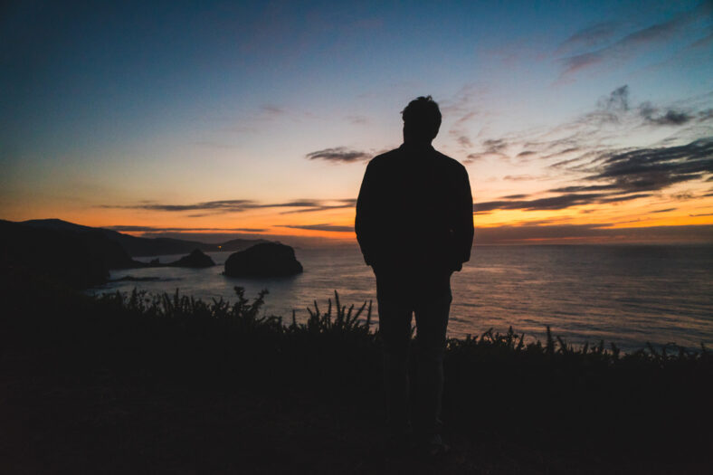 Back of a young traveler standing on on the edge of a cliff by the ocean coast watching a beautiful sunset. Freedom and wanderlust concept