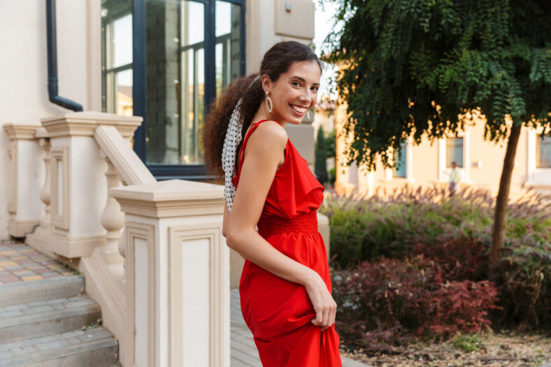 Image of beautiful young woman in red dress looking at camera and smiling while walking near building