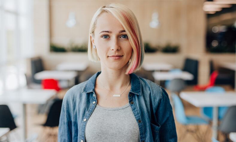 Mujer joven con rayas coloridas en el cabello mirando la cámara mientras está de pie con fondo borrosa del restaurante contemporáneo