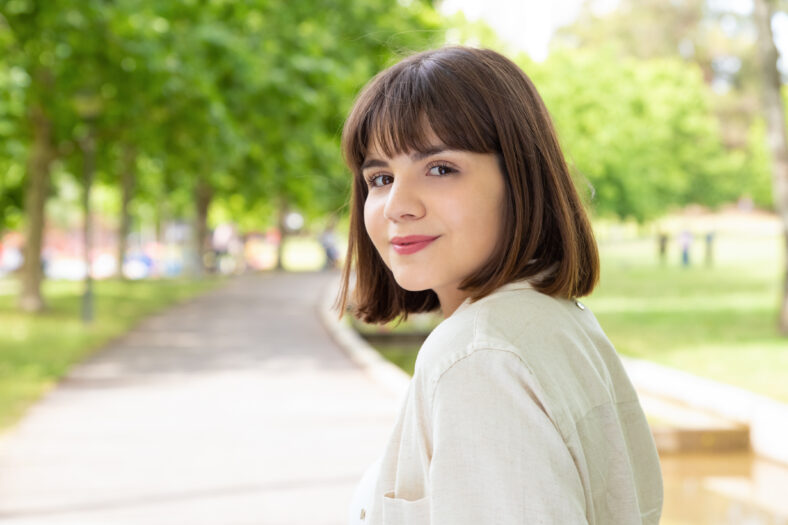 Attractive young woman smiling at camera outdoors. Portrait of beautiful cheerful young woman standing and looking at camera in park. Facial expression concept