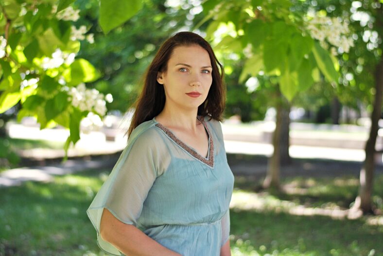 Beautiful brunette woman with long hair and blue eyes walking in the blooming park at spring in sunny day with bright sunshines and green trees
