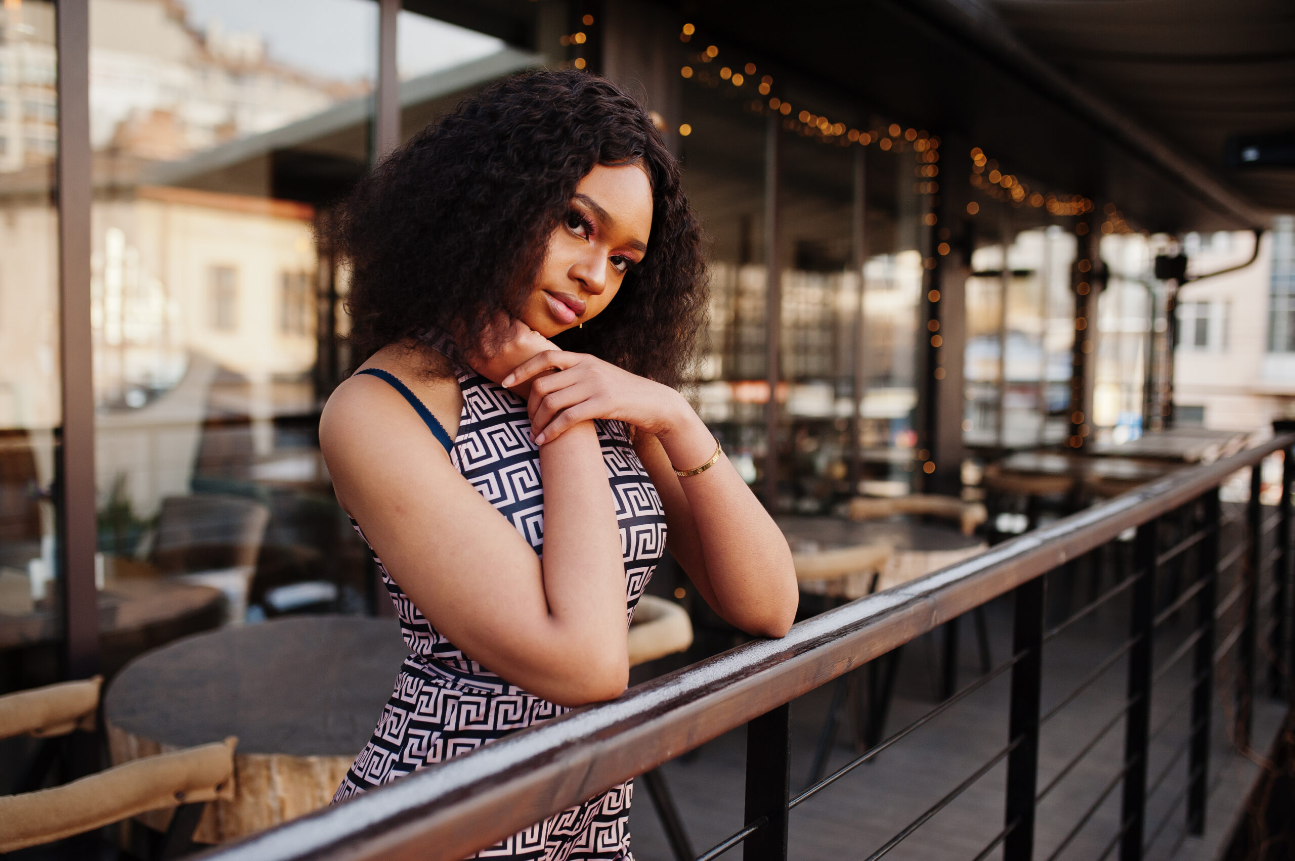 charming elegant young African american woman with long curly hair wearing jumpsuit posing at cafe terrace outdoor 