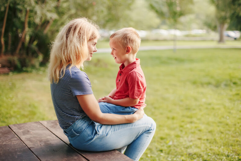 Young Caucasian mother and boy toddler son sitting together face to face. Family mom and child talking communicating outdoor on summer day. Happy authentic family childhood lifestyle.