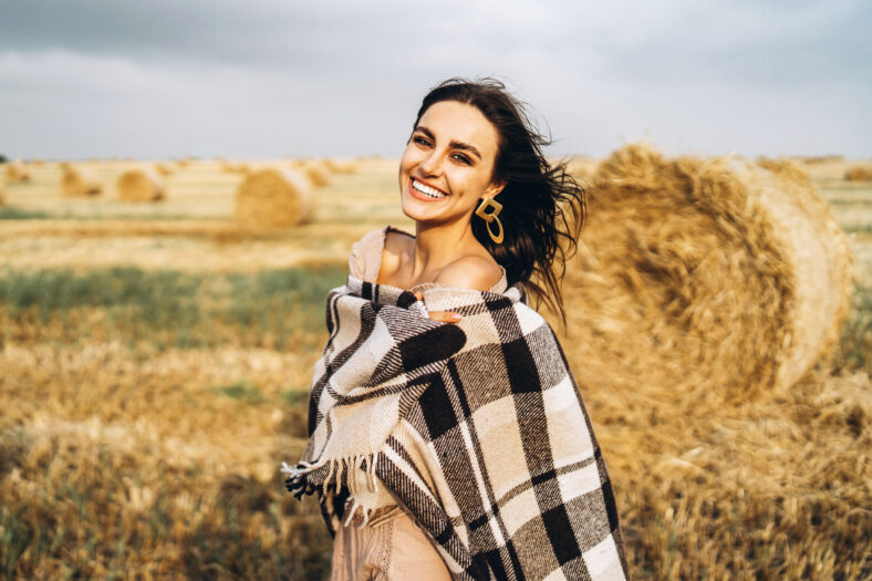 Portrait of a beautiful brunette in a dress and with a warm plaid. Woman enjoying a walk in a wheat field with hay bales.