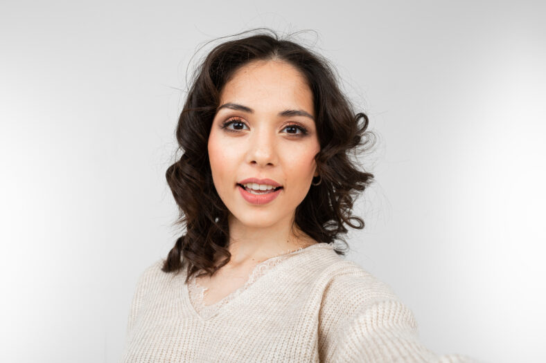La hermosa chica morena con maquillaje se hace selfie en un fondo de estudio gris.