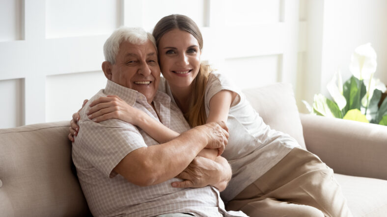 Portrait happy older father and adult daughter hugging, looking at camera, posing for photo, beautiful young woman with smiling mature dad or grandfather sitting on cozy sofa at home, two generations