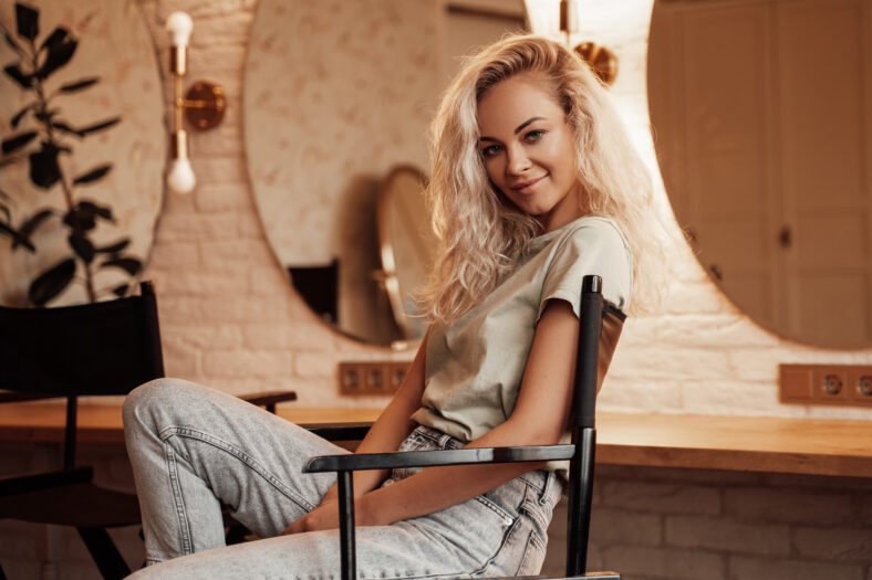 Portrait of a smiling and confident young female posing for a photoshoot in a bright room while sitting on a make up chair in front of mirrors, wearing casual clothes and white boots