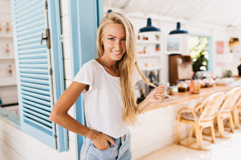 Pleasant tanned woman posing in cafeteria with hand in pocket. Indoor shot of gorgeous blonde female model expressing positive emotions during rest in restaurant