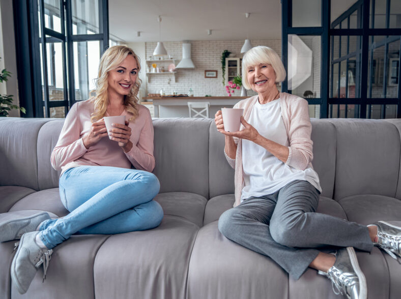 Senior woman and her attractive daughter spending time together at home. Sitting on sofa and drinking tea together. Happy Mothers' Day