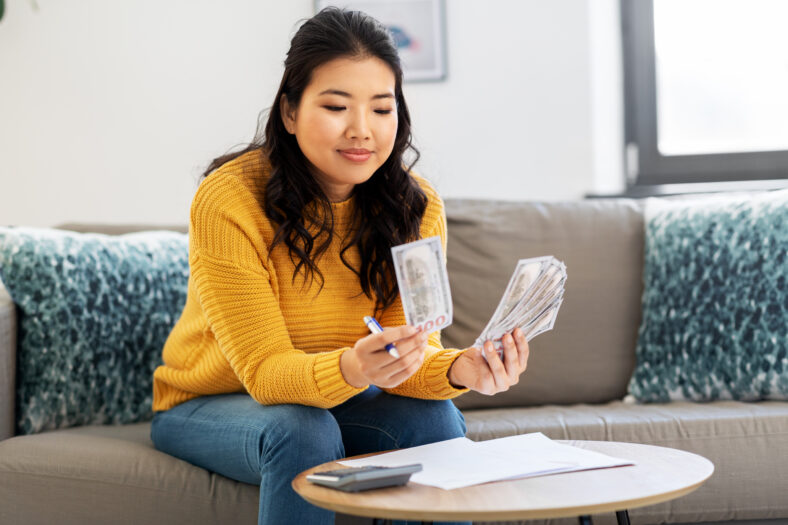 accounting, taxes and finances concept - young asian woman with papers and calculator counting money at home