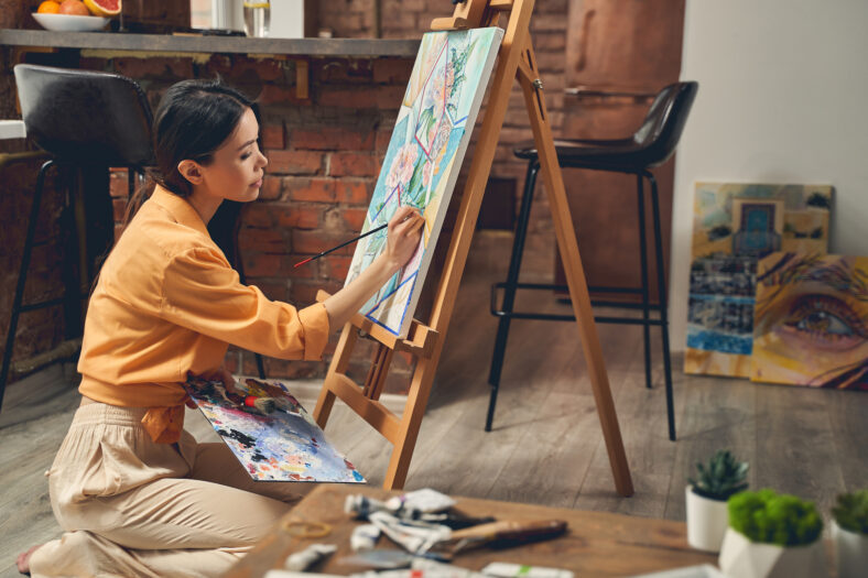 Talented female artist sitting on the floor and drawing flowers with paints on easel in home art studio