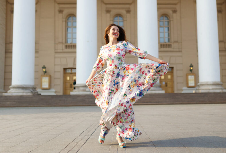 Fashion woman in spring dress on city street. Frontal view female style of feminine fashionable girl model with long wavy red hair spinning flared outdoor. Building and cobblestone pavement.