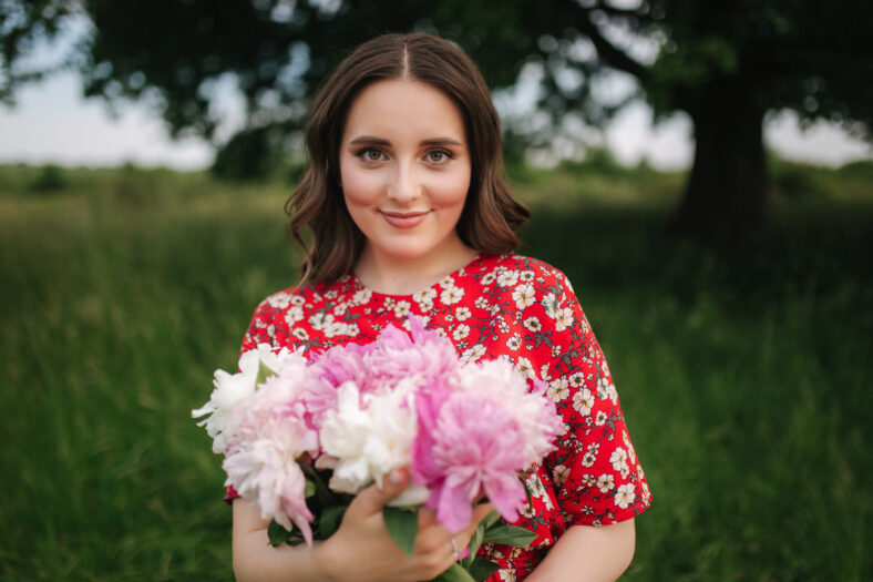 Attractive young woman in red dress with beautiful flowers walking in the field in front of big tree.