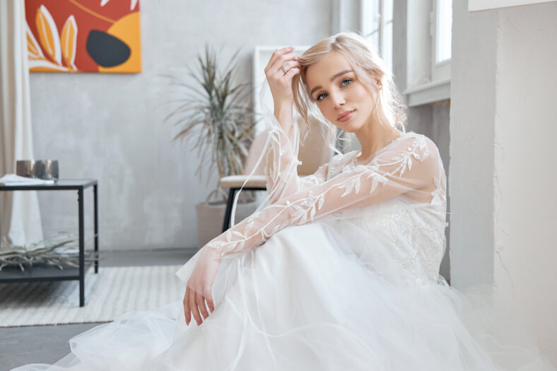 Novia ideal sentada en el piso, retrato de una niña con un largo vestido blanco. Hermoso cabello y piel limpia y suave. Peinado de boda rubio