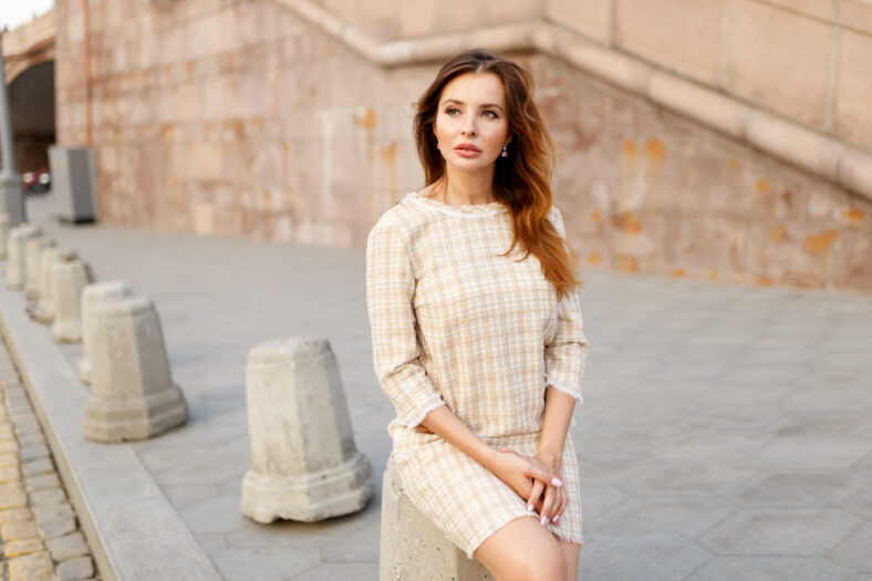 Young elegant woman stays alone in the city, dressed in a classic dress with straight and red long hair.
