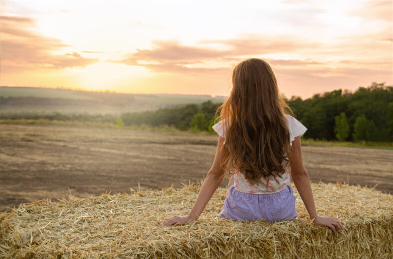 Adorable little girl child sitting on a hay rolls in a wheat field at sunset back view