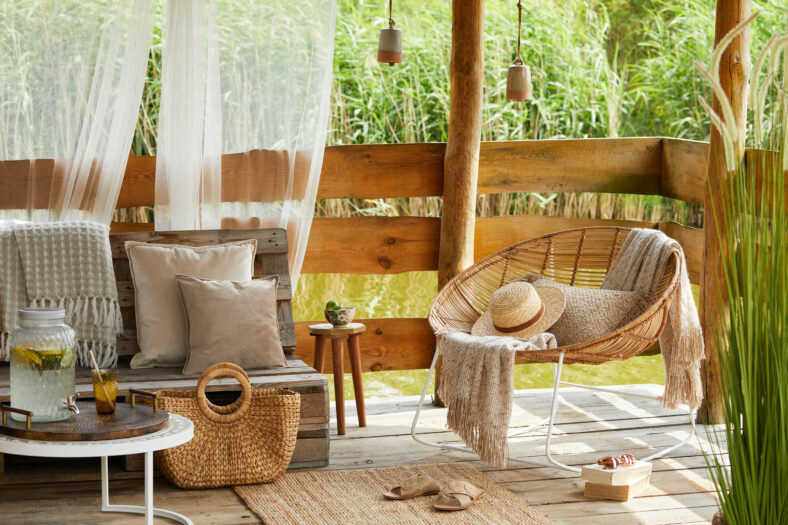Interior design of summer gazebo by the lake with stylish rattan armchair, coffee table, sofa, pillows, plaid and elegant accessories in modern decor. Summer vibes.
