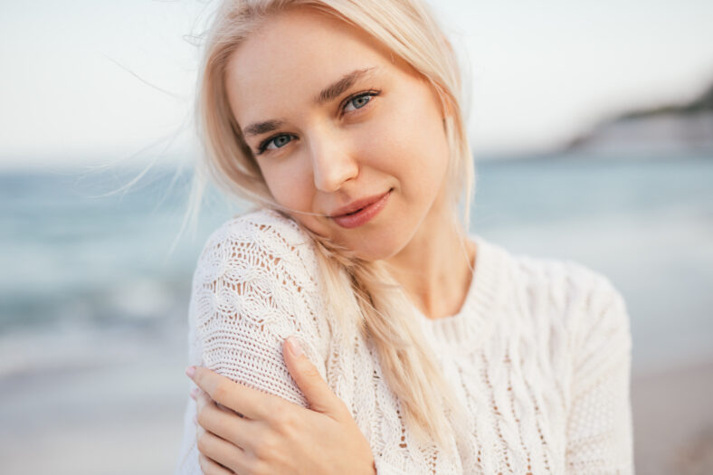Positive young blonde female in warm knitted wear smiling and looking at camera while standing against blurred sea shore