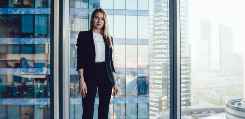 Half length portrait of pretty blonde business woman in trendy elegant formal wear standing near window in office interior, confident beautiful 30s female executive manager in suit looking at camera
